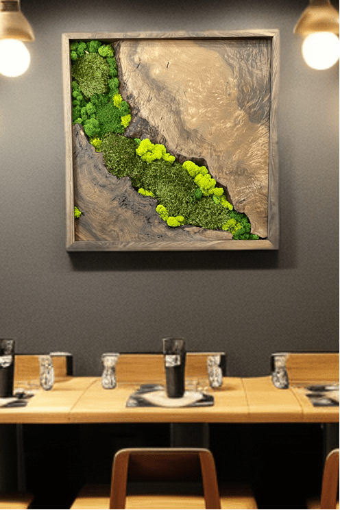 A framed square wall art piece featuring a green moss garden landscape, displayed on a dark gray wall above a wooden dining table set with chairs and place settings.