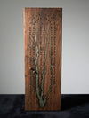 A wooden board game featuring a tree-like design made from green resin inlay, with rows of peg holes and small metal dice, standing upright on a dark surface against a light gray background.
