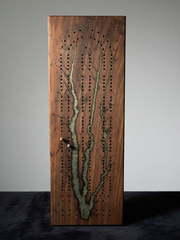 A wooden board game featuring a tree-like design made from green resin inlay, with rows of peg holes and small metal dice, standing upright on a dark surface against a light gray background.