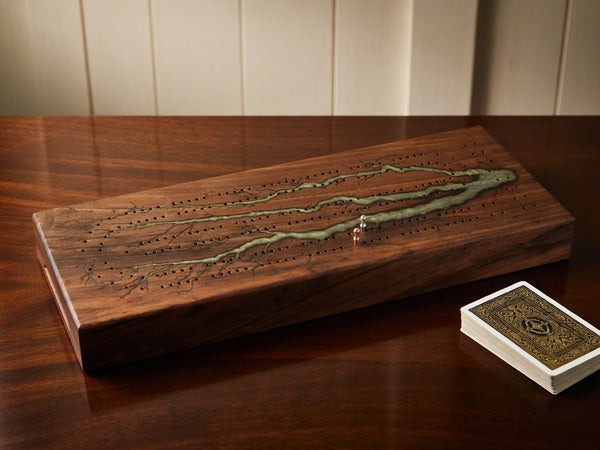 A wooden board game set displayed on a dark wood table, featuring a rectangular wooden board with engraved tracks and a green inlay, alongside a small deck of playing cards.