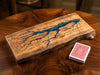 A live-edge wooden board game featuring a blue resin river running through engraved black dots, displayed on a dark wood table next to a deck of playing cards.