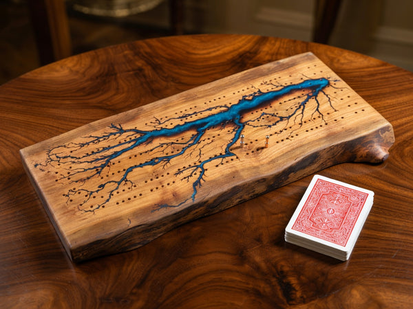 A live-edge wooden board game featuring a blue resin river running through engraved black dots, displayed on a dark wood table next to a deck of playing cards.